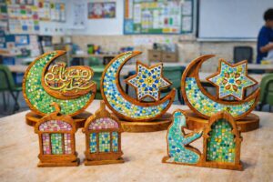 "Handmade wooden Ramadan decorations crafted with colorful mosaic tiles. Designs include a mosque, minbar, crescent moon, Umayyad star, and a girl in sujood. Each piece is displayed on a wooden surface, showcasing traditional Islamic art with a modern educational twist. Sizes vary from 20x30 cm to 35x25 cm."